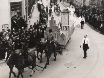 Historisches Foto: Pferde ziehen Festwagen durch eine Menschenmenge in einer Stadt.