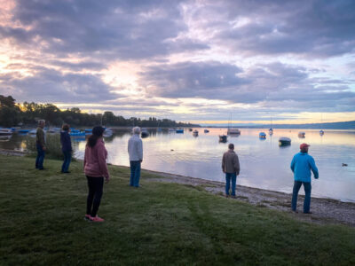 Personen am Seeufer bei Sonnenuntergang. Boote auf dem Wasser, bewölkter Himmel.